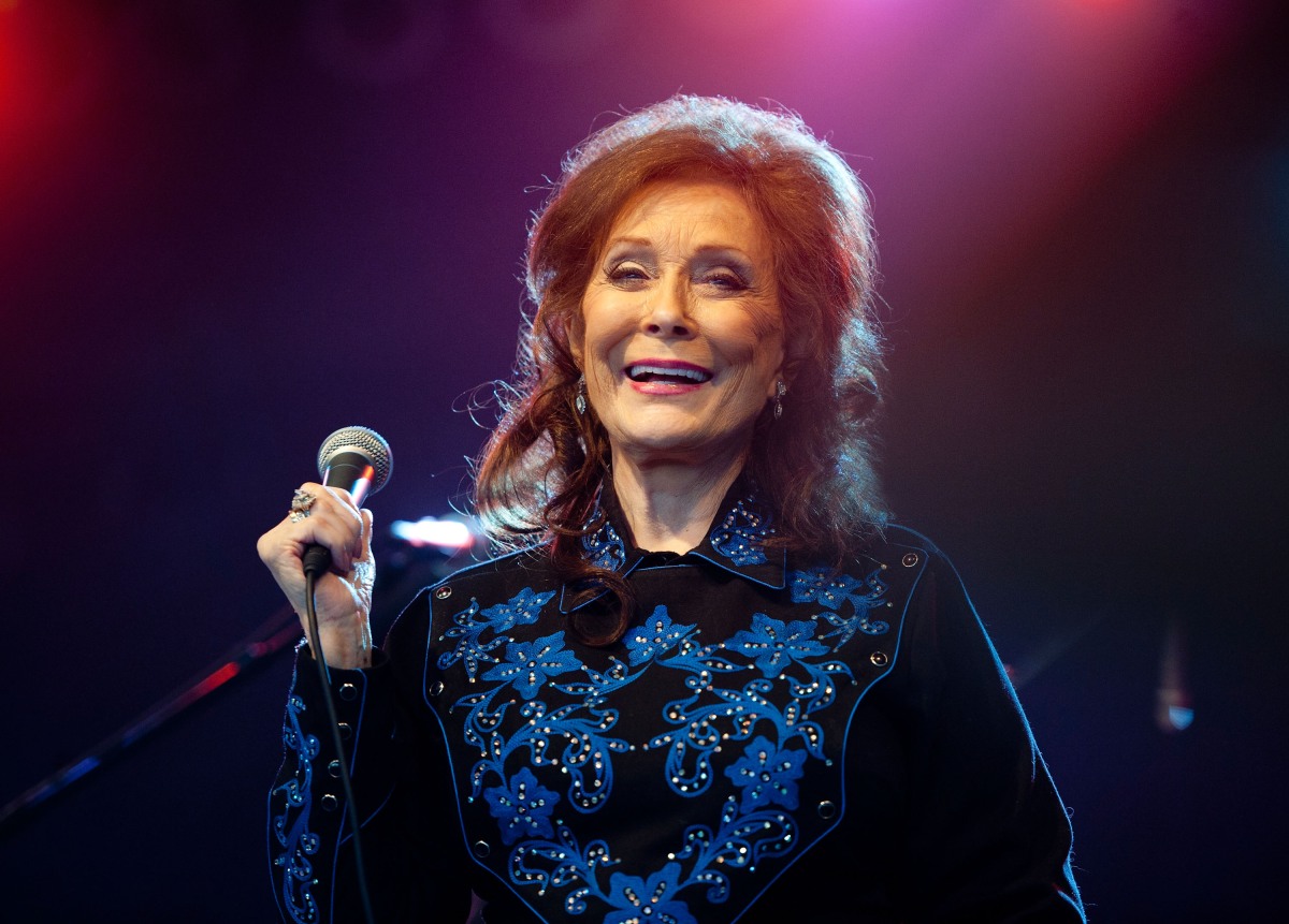 Loretta Lynn holds a microphone on stage while wearing a blue-and-black shirt